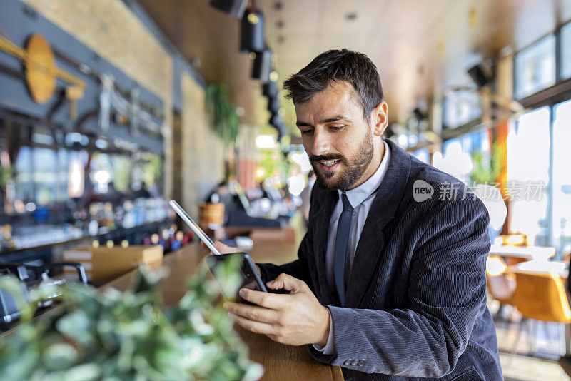 一个微笑的商人正在现代咖啡店里用手机和他的商业伙伴交流。他正坐在他的数字平板电脑，享受着一杯咖啡。