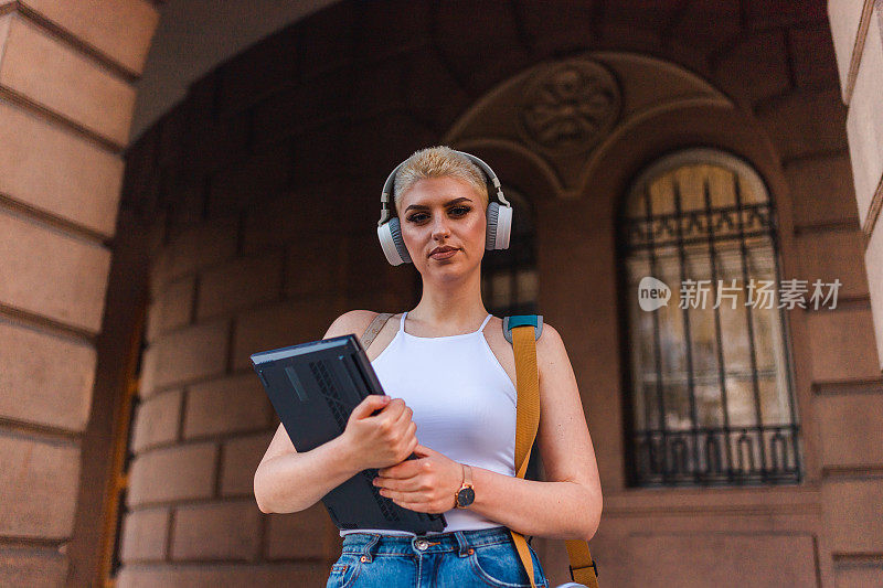 一名金发短发的年轻女学生的齐腰肖像，戴着耳机，拿着笔记本电脑