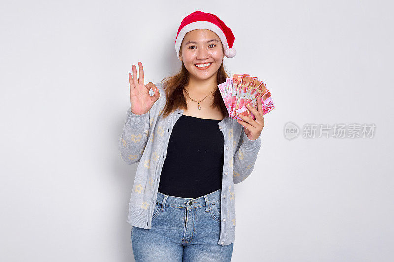 微笑的年轻亚洲女子20岁戴着圣诞帽，上面有现金、印尼盾和白色背景上的okay标志