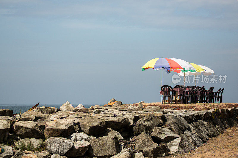 在海边的沙滩上，五颜六色的阳伞下摆放着一组桌椅