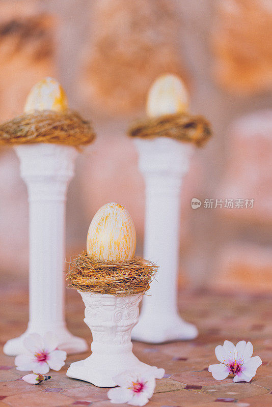 石墙前的一张旧桌子上有三个金色的复活节彩蛋，上面有白色的小柱子和杏仁花