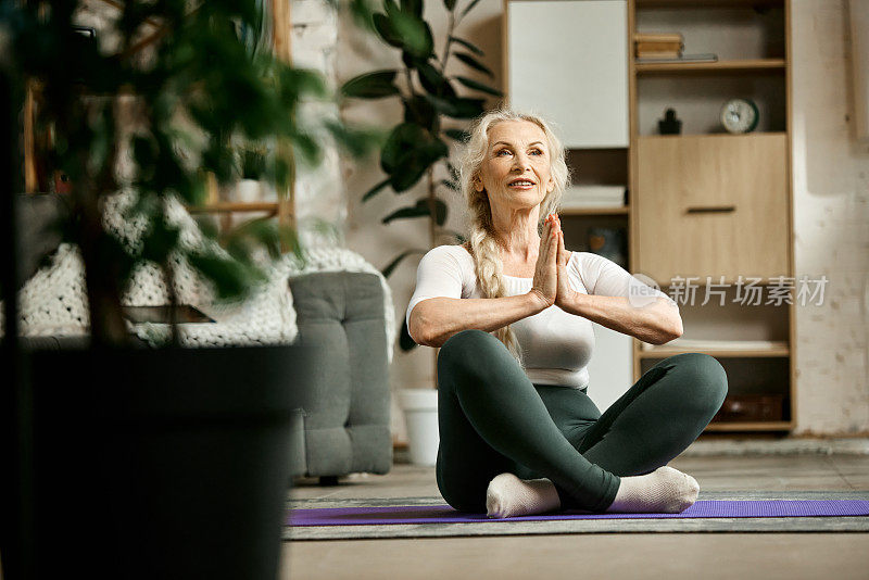 美丽，积极，高级女性坐在客厅的健身matt上，在家做瑜伽练习。冥想