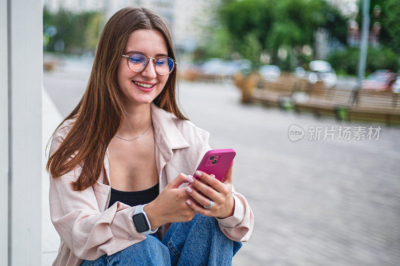 一个穿着牛仔裤、衬衫、拿着手机的年轻女孩坐在街上微笑着
