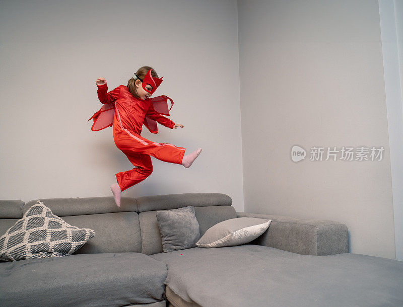可爱的小女孩穿着红色的超级英雄服在公寓里跑来跑去，蹦蹦跳跳，在半空中飞翔