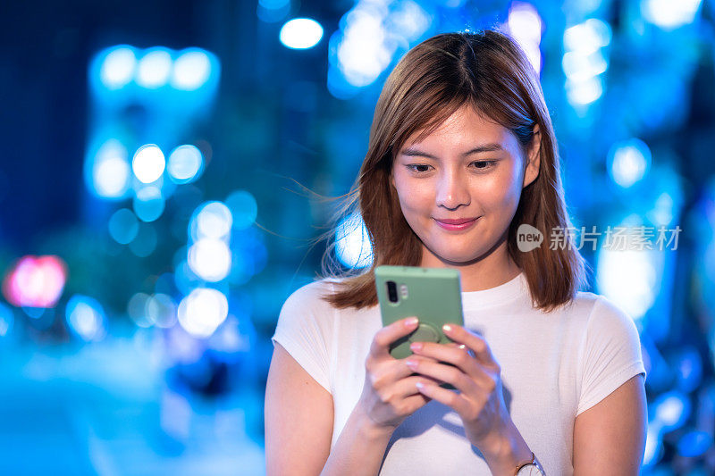 年轻漂亮的亚洲女人晚上在城市里用手机，背景是建筑散景灯