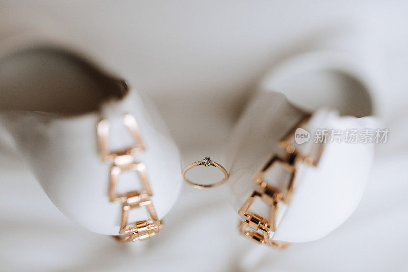 一枚优雅的镶有钻石的金婚戒，立在女士们的白色高跟鞋之间。上图为白色背景