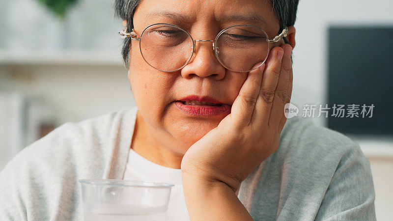 苦恼的亚洲老人坐在家里的客厅里抚摸着牙齿问题的脸颊、牙齿敏感的祖母、牙齿疼痛的老年妇女