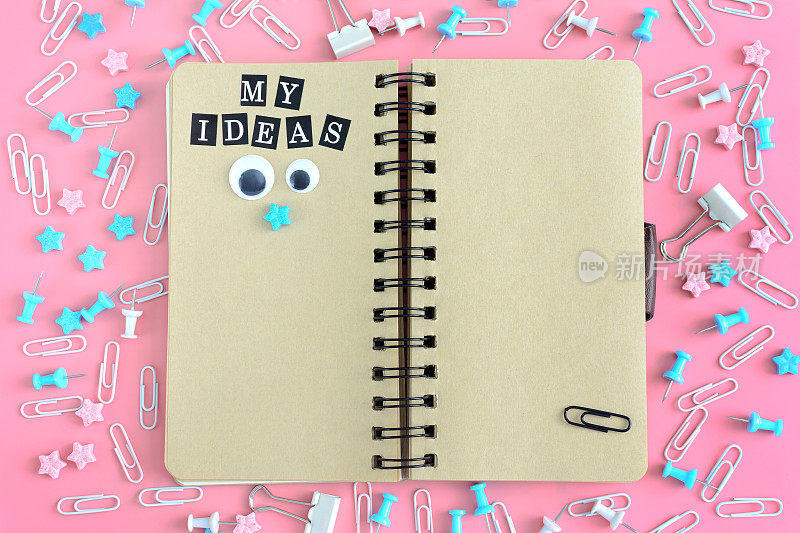 弹簧上的记事本是打开的。在文具旁边。在笔记本棕色的书页上写着“我的想法”。上面的照片，粉红色的背景。