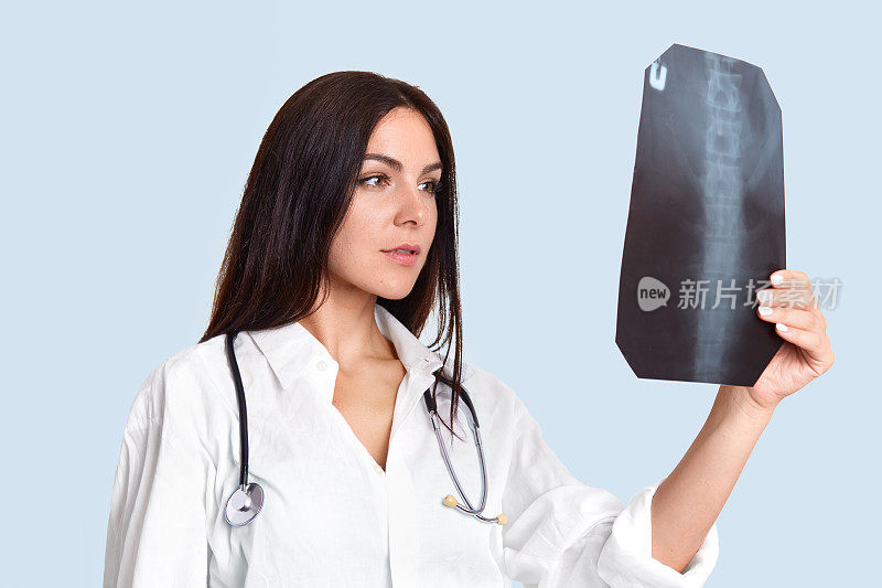 严肃的女医生仔细地看X光片，检查人们的脊柱，穿着白色的实验服，脖子上有听诊器，在浅蓝色的背景下侧身站立。医疗的概念