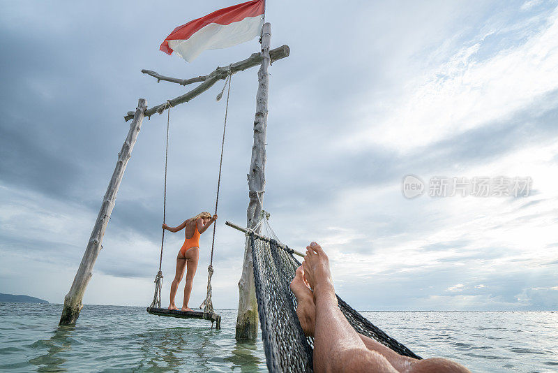 年轻夫妇在海边的海滩上荡秋千，美丽的田园诗般的风景。人们旅行浪漫度假的概念。男人在吊床上和女朋友在海上荡秋千的个人观点。