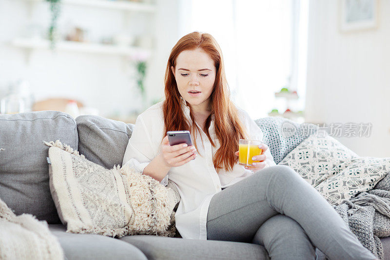 红头发的漂亮年轻女子坐在家里的沙发上，用手机浏览互联网，看起来很惊讶。她手里拿着一杯橙汁