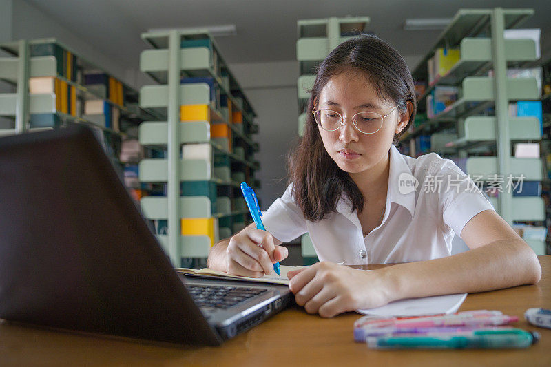 亚洲大学生女孩读书和工作用笔记本电脑和智能手机做她的家庭作业在公共图书馆