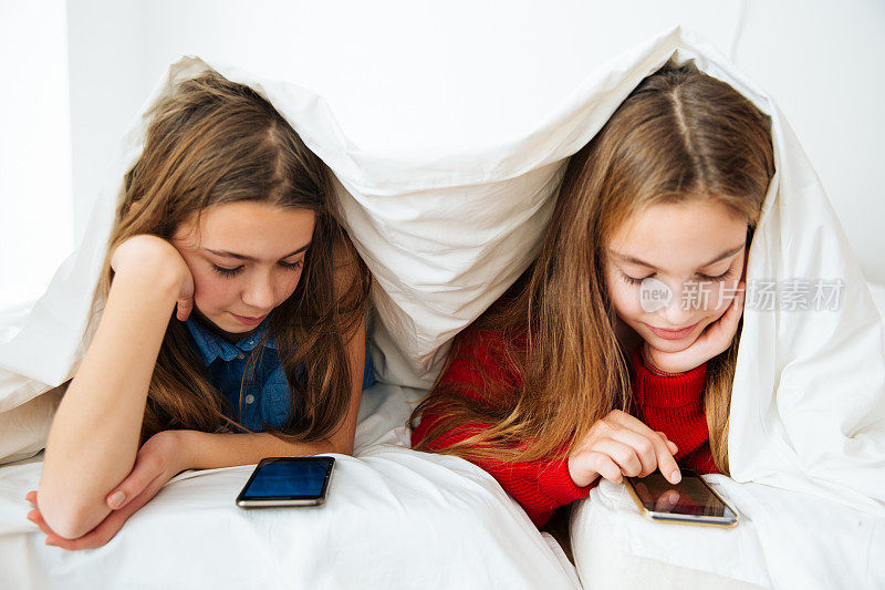 两个小女孩躺在毯子下看手机