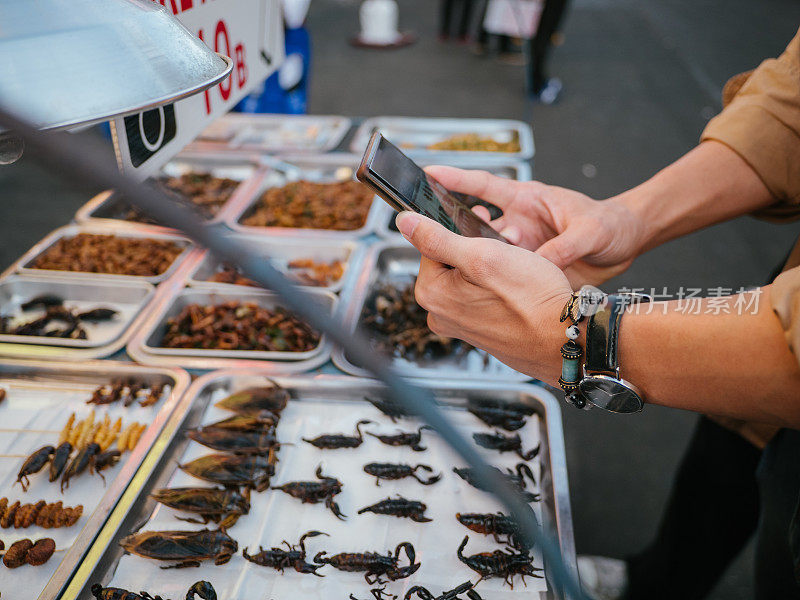 一名亚洲男子在街头美食旁拍摄被炒昆虫。曼谷,泰国。