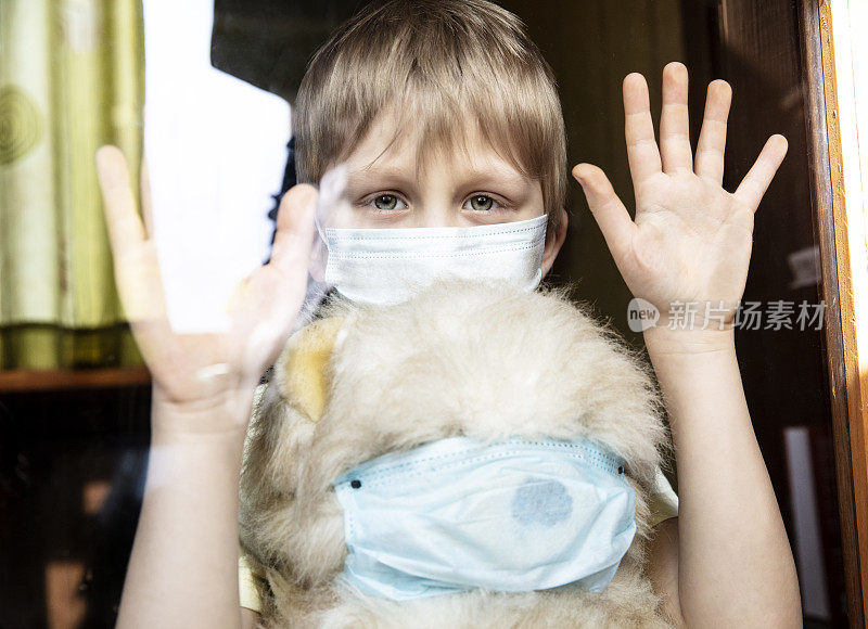 一个男孩带着他生病的泰迪熊，戴着医用防护口罩从窗户往外看。预防流行病。