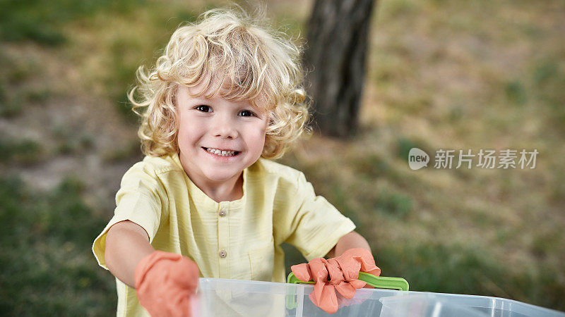 快乐的小男孩戴着橡胶手套，拿着回收箱，微笑着对着镜头，与父母在森林或公园收集塑料垃圾
