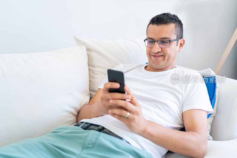 躺在沙发上用智能手机的快乐微笑的男人。在家放松