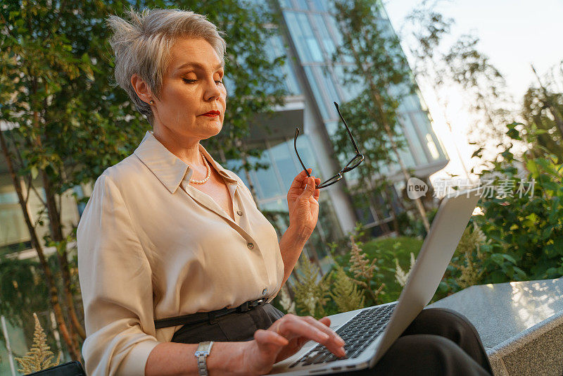 这是一位身穿古典服装、头发灰白的商界女性的低视角照片，她正看着自己的笔记本电脑，手里拿着眼镜