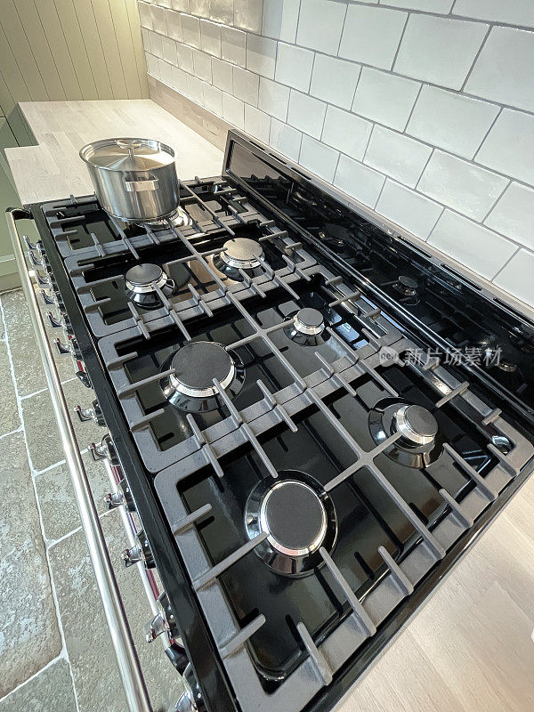 近景现代厨房7燃烧器煤气灶在轻木效果台面，橱柜照明下，黑色玻璃表面，排表盘和旋钮，不锈钢烹饪锅，高架视野，聚焦前景
