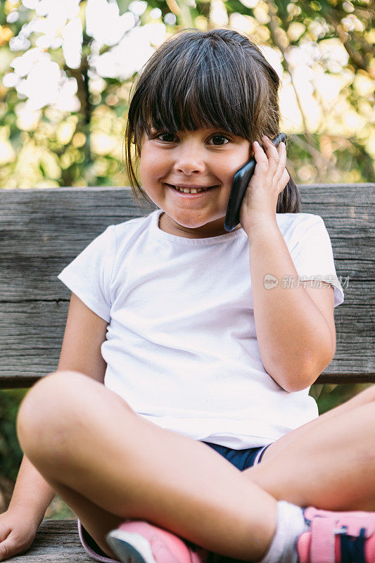 黑头发的小女孩，穿着白色t恤，坐在公园的长凳上，微笑着讲手机。