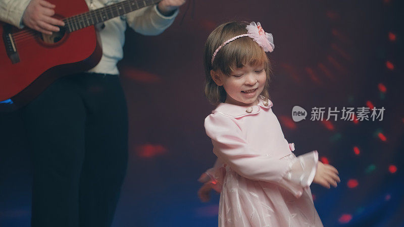 穿着复古礼服的小女孩在舞台上跳舞，她的父亲弹着原声吉他
