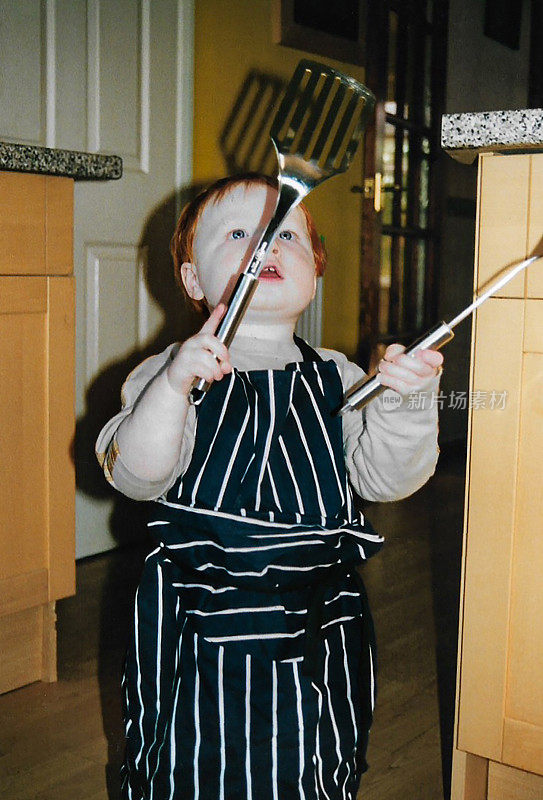 一个红头发的小男孩在家庭厨房里拿着一个金属铲和勺子，穿着超大的屠夫围裙，聚焦在前景上