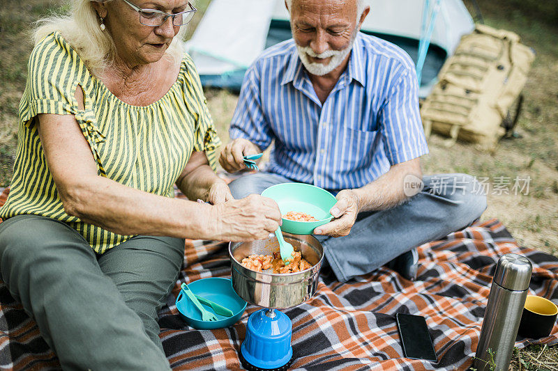 一对老年夫妇在户外露营时烹饪和制作食物。夏日的露营地，一男一女坐在帐篷外。