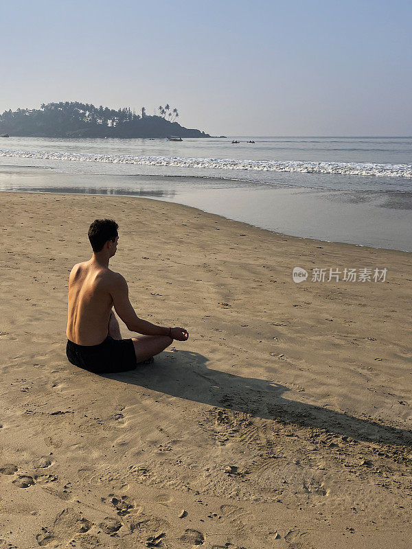 裸胸印度男子在潮湿的沙滩上练习莲花姿势(莲花姿势)瑜伽的图像，水的边缘，专注于前景，后视图