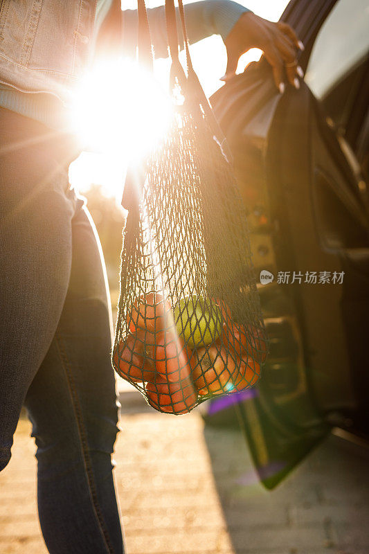 一个年轻的黑人妇女打开她的汽车后门，把苹果装在一个可重复使用的网袋里