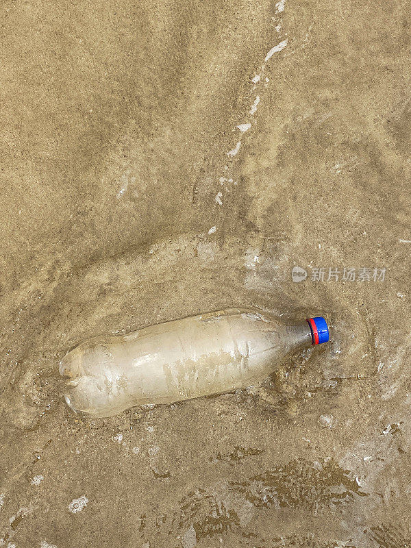 图片:空的，透明的塑料饮料瓶，蓝色盖子被冲到退潮水边缘的沙滩上，海浪冲刷着海洋垃圾和岸上的污染，乱扔的沙子，肮脏的海滩，高架视图，复制空间