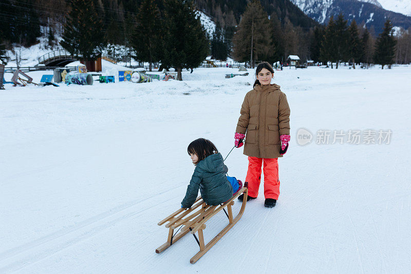 大一点的孩子和小一点的孩子一起拉雪橇，背景是美丽的冬天的山