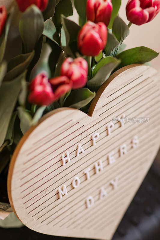 一个心形的字母板，上面写着“母亲节快乐”，明亮的粉红色郁金香盛开，郁郁葱葱的绿色茎和叶子在它后面，在明亮的自然日光下拍摄