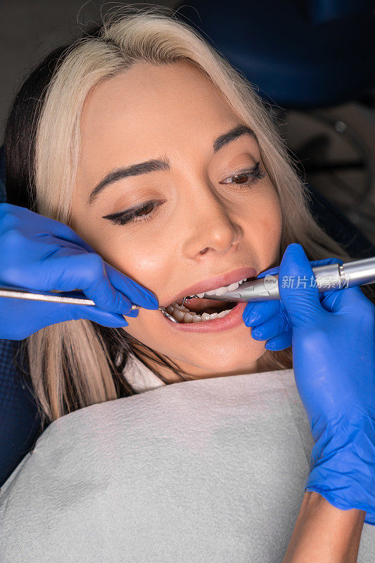 牙医在牙科椅上给女病人钻牙。牙科医生手握器械在病人口腔内进行龋齿治疗的特写。垂直图