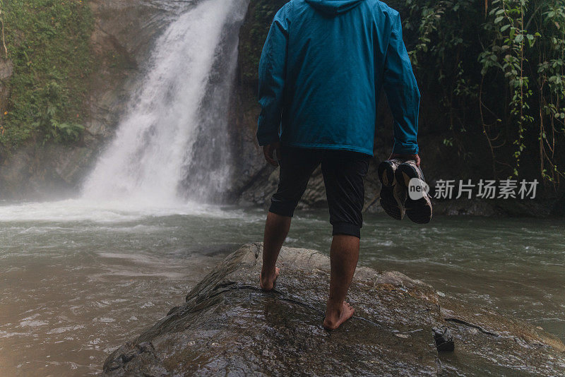 一名亚洲男子在热带雨林中拿着鞋子欣赏瀑布美景