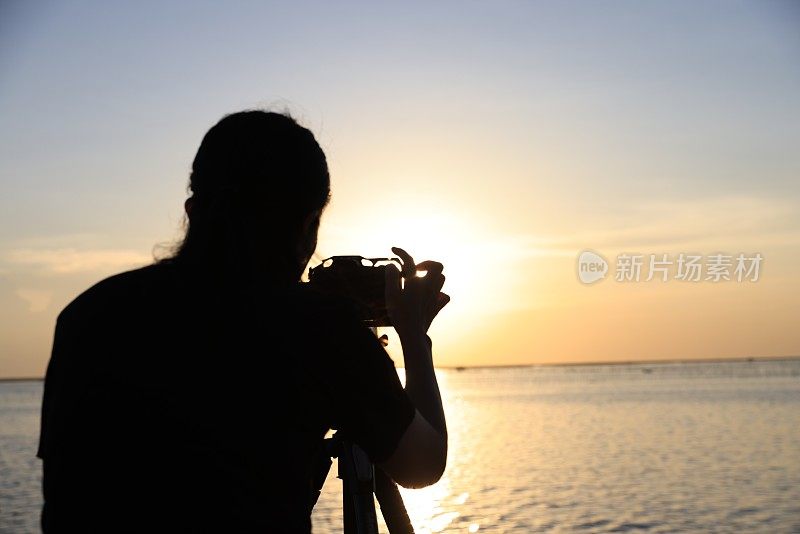 一个女人在夕阳下拍照的剪影。摄影师用剪影在桥上拍摄日落照片。准备好相机和三脚架，准备在桥上拍一张日落照片。