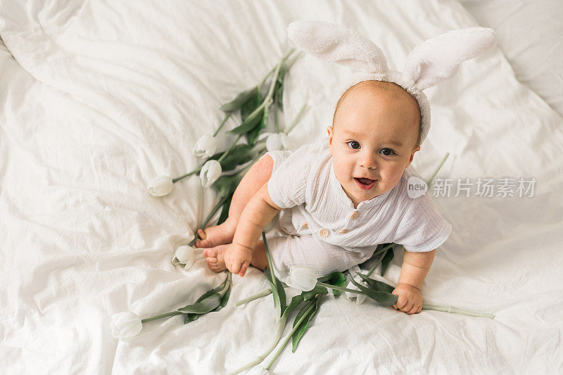 可爱的宝宝头上戴着兔子耳朵，家里的床上放着郁金香。复活节和春天时间的概念。