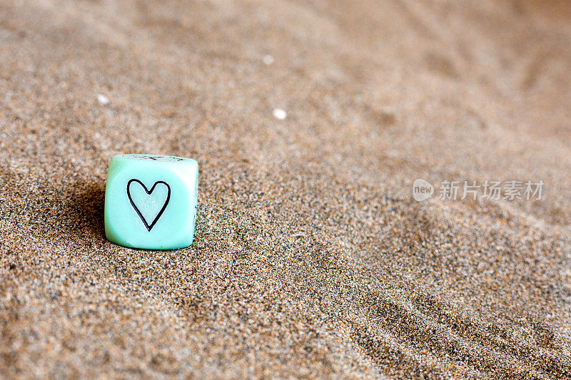 浅蓝色立方体，侧面有心形和天气符号。沙子上有趣的赌博骰子。情人节的概念。爱的象征。带有复制空间的特写图片。柔情表达