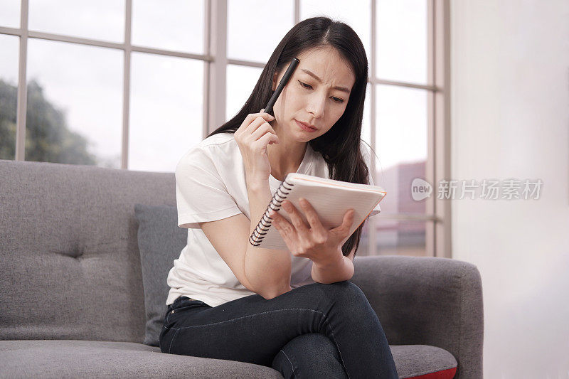 严肃的亚洲女人正在笔记本上思考和写作。她坐在办公室的沙发上微笑着学习业务。女商人的生活方式和作家的概念