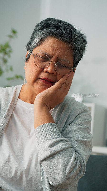 有牙齿问题的亚洲老年妇女坐在家里的沙发上抚摸脸颊、牙齿敏感的不快乐的祖母、牙齿疼痛的老年妇女