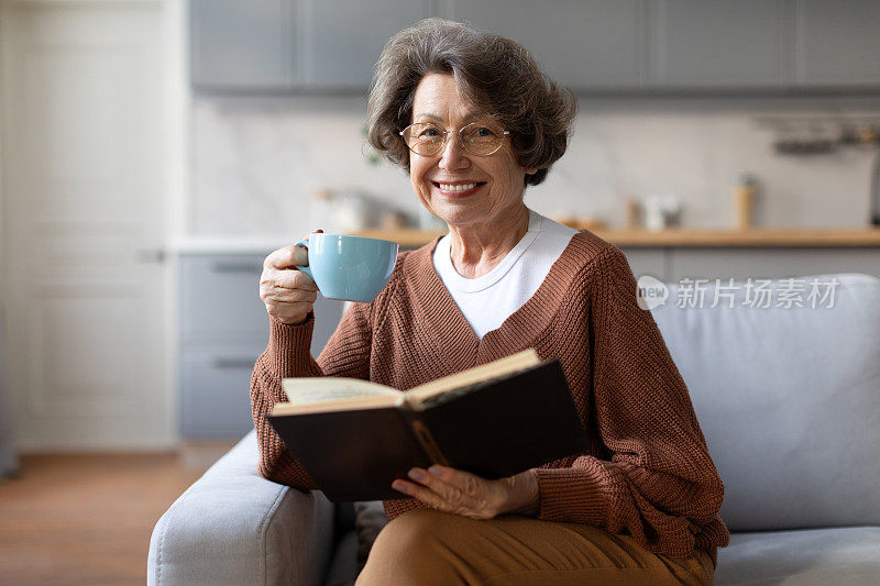 快乐的老年妇女喝着热茶，读着喜欢的纸质书，享受惬意的周末，独自坐在沙发上放松，对着镜头微笑