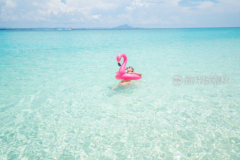 年轻女子放松在田园诗般的海滩充气火烈鸟玩在纯净的水在泰国岛屿。人们旅行奢侈，乐趣和酷的态度概念