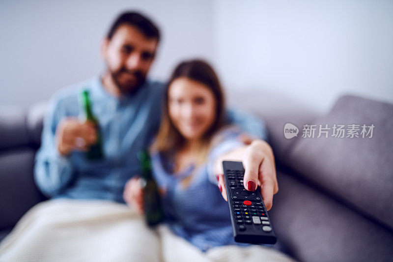 一对漂亮的白人夫妇盖着毯子坐在客厅里看电视喝啤酒。拿着遥控器换频道的女人。选择性的专注于远程控制。