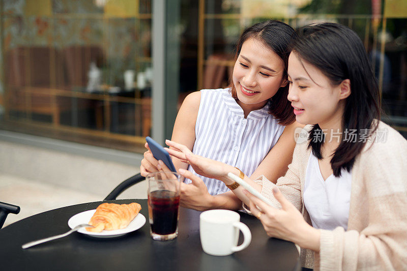 两个年轻女人一起喝咖啡休息时间用智能手机。在路边咖啡馆用手机的快乐女人