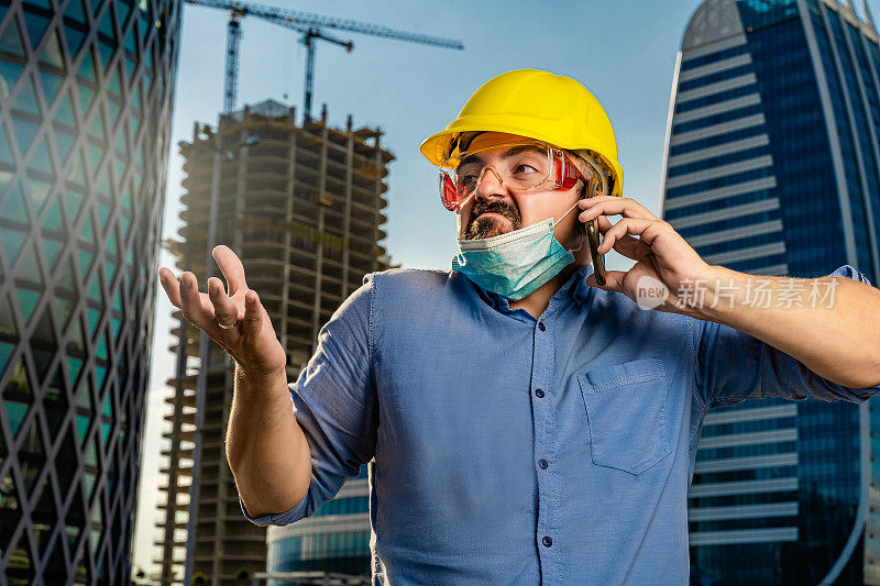 年轻工程师戴着黄色安全帽和N95防护面具在现代商业建筑和建筑工地前使用智能手机
