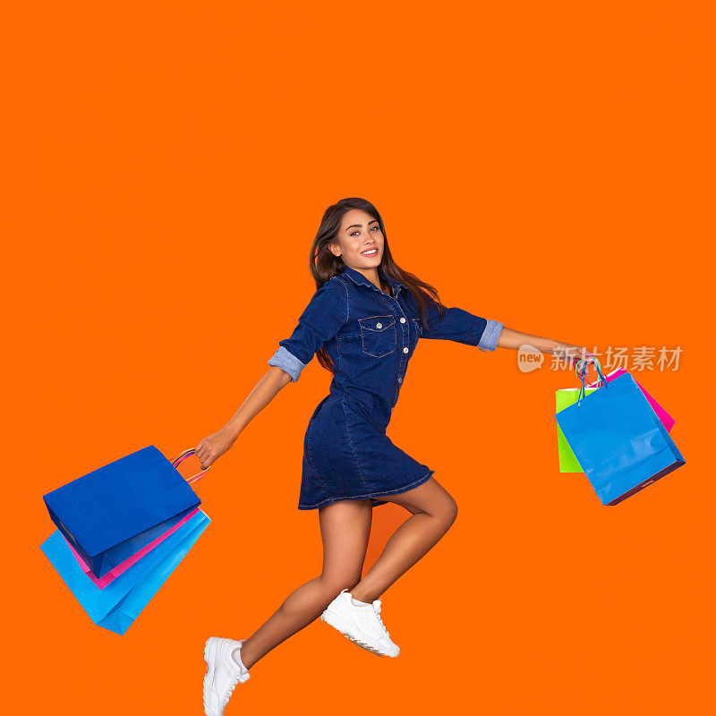 活泼的年轻女子穿着牛仔裙，橙色背景上的购物袋在空中。购物概念，夏季大甩卖