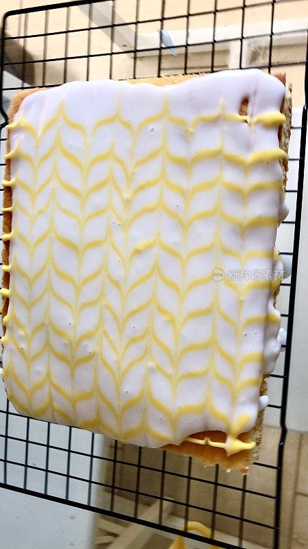 这是一个未经切割的自制柠檬蛋糕，上面覆盖着白色翻糖糖霜和羽毛状的黄色柠檬味糖霜，放在金属冷却架上，居高临下