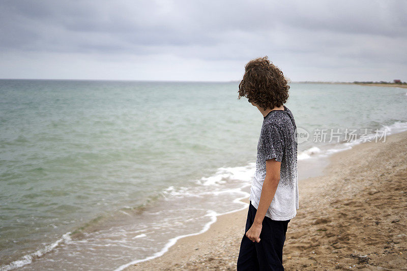 在一个多云的夏日，一个留着卷发的15岁男孩独自沿着海边的海滩走着，望着远方的地平线。