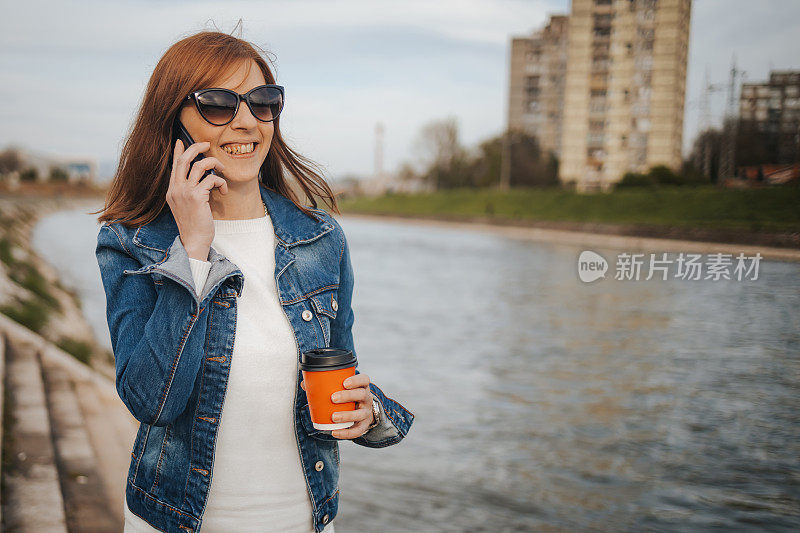 一个年轻女子在城市码头边打电话边喝外卖咖啡