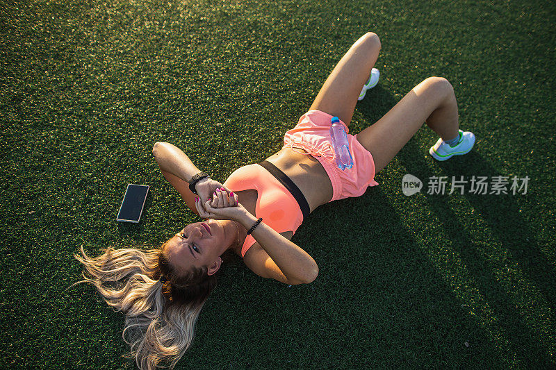 健身女孩穿着运动短裤和上衣休息在体育场的草地上。检查smartwatch。训练和休息的概念