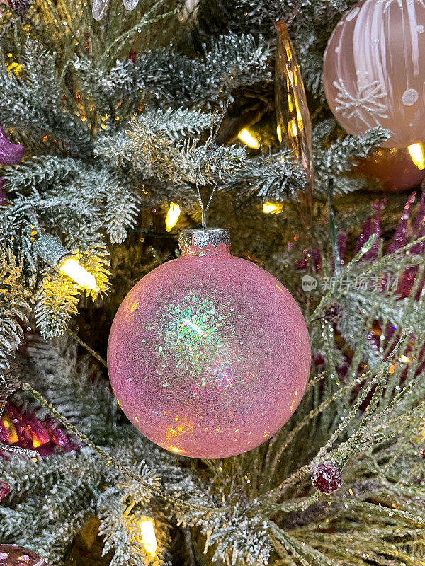 个人的图像，粉红色的闪光小饰品挂在人造圣诞树的树枝上，云杉针覆盖着人造喷雾雪，模糊的白色精灵灯的背景